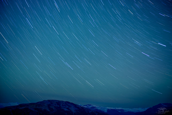 Rocky and snowy mountains are at the bottom of the image. The rest of the image is filled with star trails. The background is blue, yellow and green. Fine art photography by Shel Neufeld, wildlife and nature photographer of Robert's Creek, BC, Canada. 