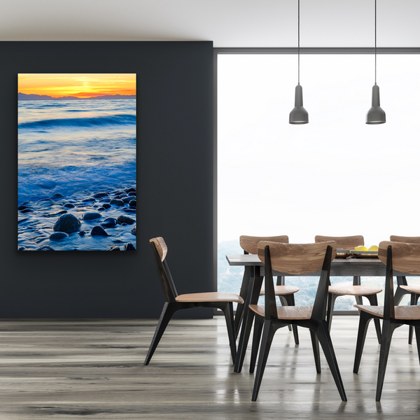 A coastal sunset with waves and a rocky beach are captured by Shel Neufeld. Available for purchase in a variety of sizes and print mediums. The large vertical wall art hangs in a dining room. 
