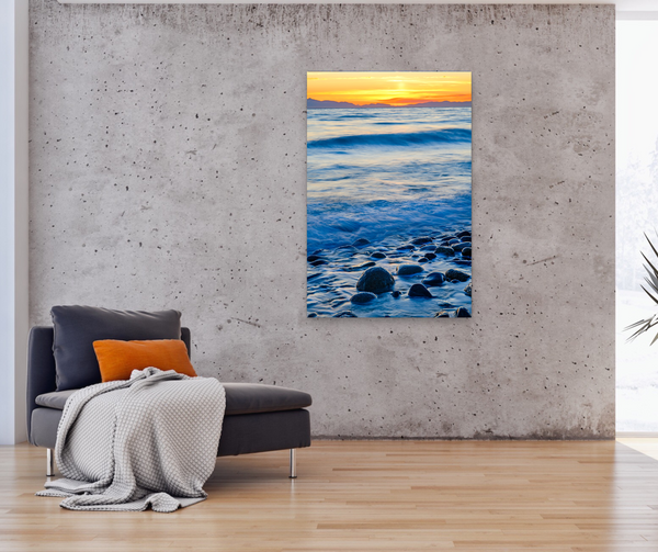 A coastal sunset with waves and a rocky beach are captured by Shel Neufeld. Available for purchase in a variety of sizes and print mediums. The large vertical artwork hangs in a living room. 