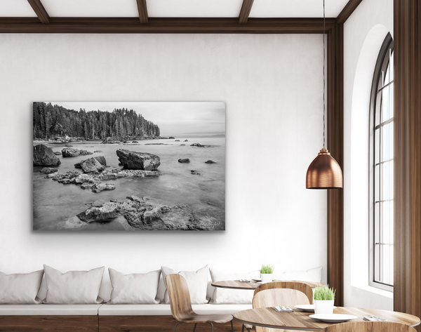 A black and white photography canvas of Sombrio Beach is hung on the wall above a wall couch. To the right of the artwork is a large window and a small table with setting for two. This image is used to visualize the photography canvas within a home space.  The photography canvas print is available for sale. Artwork by Shel Neufeld from Roberts Creek, BC, Canada. 
