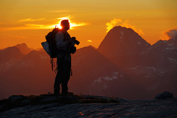 Photographer Shel Neufeld stands on a mountain ridge with his camera in hand and a backpack on. The sun sets behind him, lighting the sky with orange. 