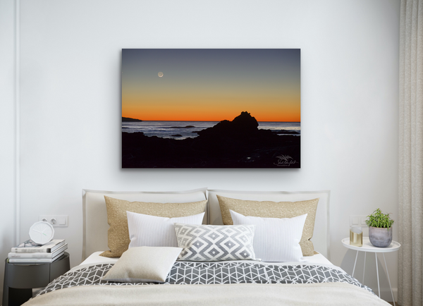 A photography canvas print of Sombrio Beach sunset with the moon overhead captured by Shel Neufeld. The canvas hangs in a bedroom scene. 
