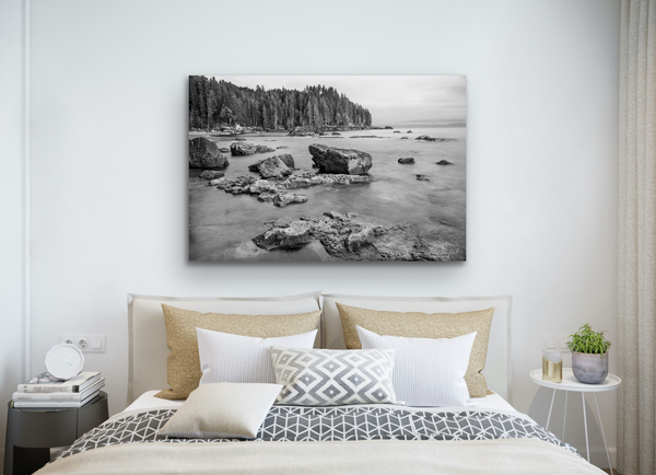 A black and white photography canvas of Sombrio Beach is hung on the wall above a bed. The bed is filled with white, beige and grey bedding. The canvas is 40x60 and is available for purchase. The photography canvas print is available for sale. Artwork by Shel Neufeld from Roberts Creek, BC, Canada. 