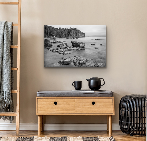 A black and white photography canvas of Sombrio Beach is hung on the wall above a tea set and magazine. To the left of the canvas artwork is a wooden decorative ladder and a grey throw blanket. To the right is a metal ottoman. The photography canvas print is available for sale. Artwork by Shel Neufeld from Roberts Creek, BC, Canada. 