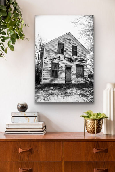 Black and white vertical photography canvas hangs in a living room. It contains a picture of an abandoned house. Artwork by Shel Neufeld, Canadian landscape and nature photographer. 