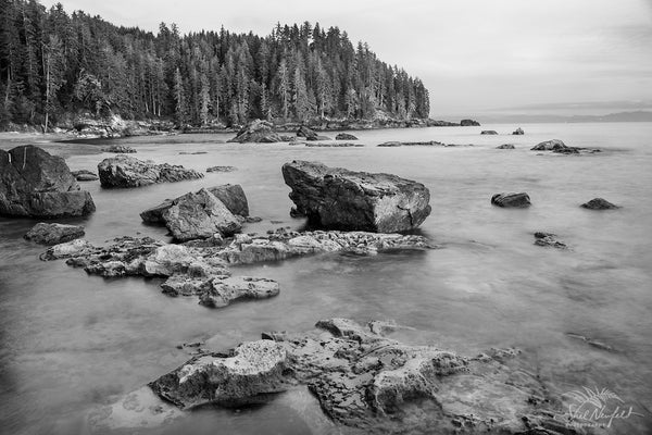 Sombrio Beach captured in black and white and with a long time exposure. The scene contains a rocky shore with smooth water around the rocks. In the distance is a piece of land extending into the ocean filled with tall trees. In the distance you can see more land. This photograph is captured by Canadian landscape artist, Shel Neufeld of Roberts Creek, BC, Canada. 