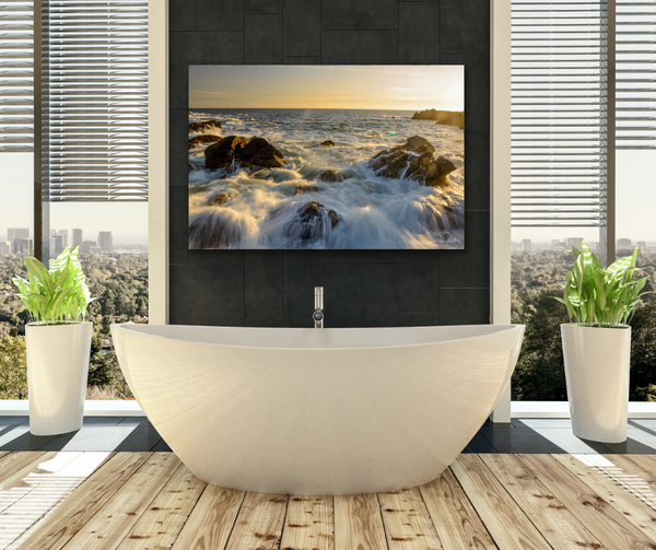 Ucluelet Water crashing large wall art photography canvas. The canvas hangs on the wall of a bathroom decor. The bathroom scene contains the large ocean landscape canvas, a big white soaker bathtub, two large floor to ceiling windows, green plants in white planters and a rustic wood floor. The image is to help visualize the photography canvas in a bathroom decor. Artwork by Shel Neufeld of Roberts Creek, BC, Canada. 