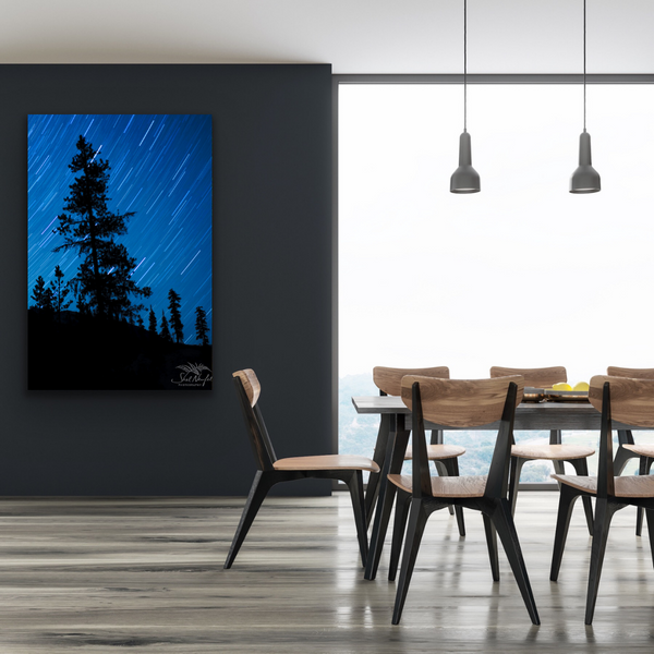 A large vertical wall art hangs on the grey wall in the left of this image. The wall art is a photograph of a big night sky with stars and tree silhouettes in the foreground. The large vertical wall art is in a living room scene with modern wood and black chairs and a large picnic style table. It is in front of a large window. Artwork by Shel Neufeld, Canadian landscape and nature photographer