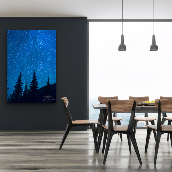 A big starry night canvas with tree silhouettes in the foreground. The artwork is by Canadian landscape and nature photographer Shel Neufeld. The print hangs in a dining room scene with wooden chairs, a large picnic style table and a large window. 