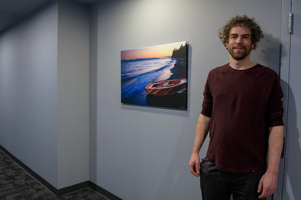 Photographer Shel Neufeld stands beside one of his photography canvas wall art pieces that hangs in a building. 
