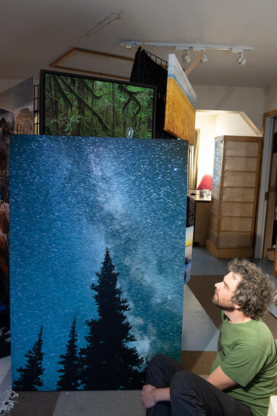 Photographer Shel Neufeld sits in front of a large photography canvas of his. This image is milky way starry night. 
