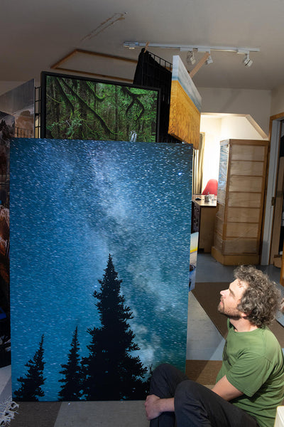 Photographer Shel Neufeld sits in front of a large canvas print of his photography. The large canvas measures 40 inches by 60 inches. It is his milky way starry night image. 